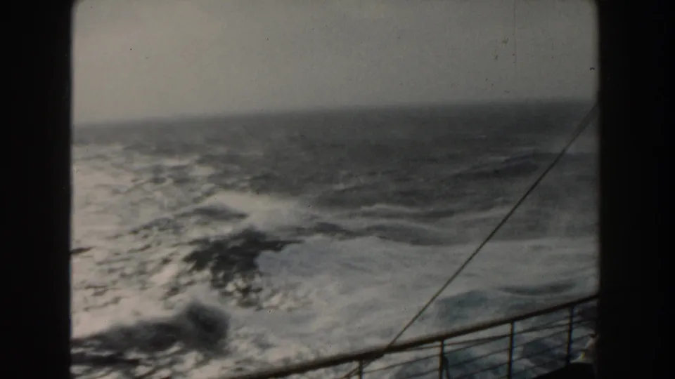 A deck-level 8mm frame shows rolling open ocean, whitecaps, a low horizon, and the ship railing in the lower foreground.