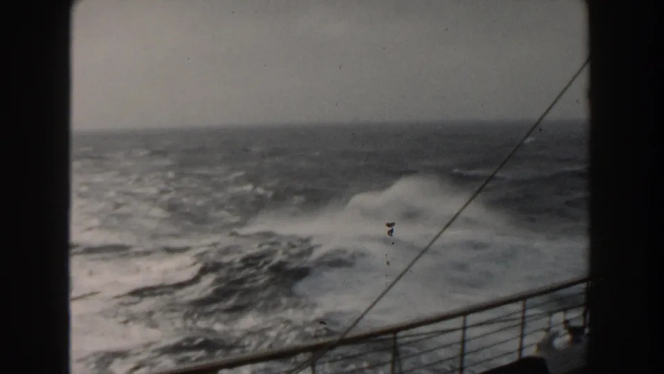 A burst of ocean spray rises near the ship railing, with rough Atlantic water, a diagonal cable, and faded 8mm texture visible.