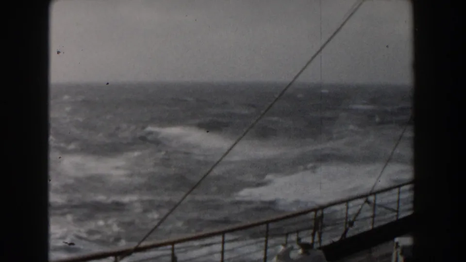 Shipboard 8mm frame showing rough Atlantic water, whitecaps, deck railing, and rigging lines in stormy conditions.