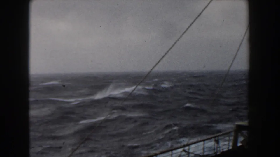Rough Atlantic waves and spray fill the frame, with ship railing and rigging visible along the lower and right edges.