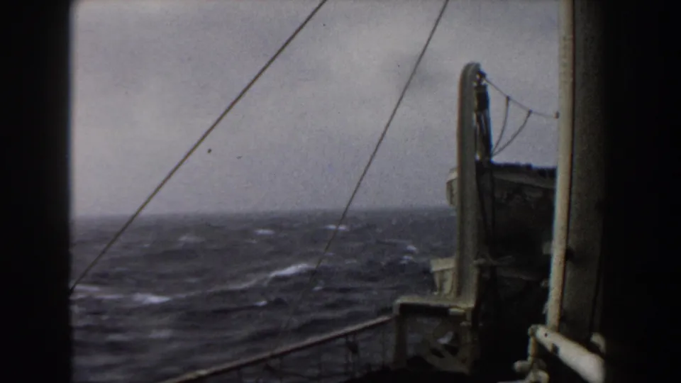 Shipboard 8mm view of choppy Atlantic water, gray horizon, deck rail, rigging, and a vertical ship structure at frame right.