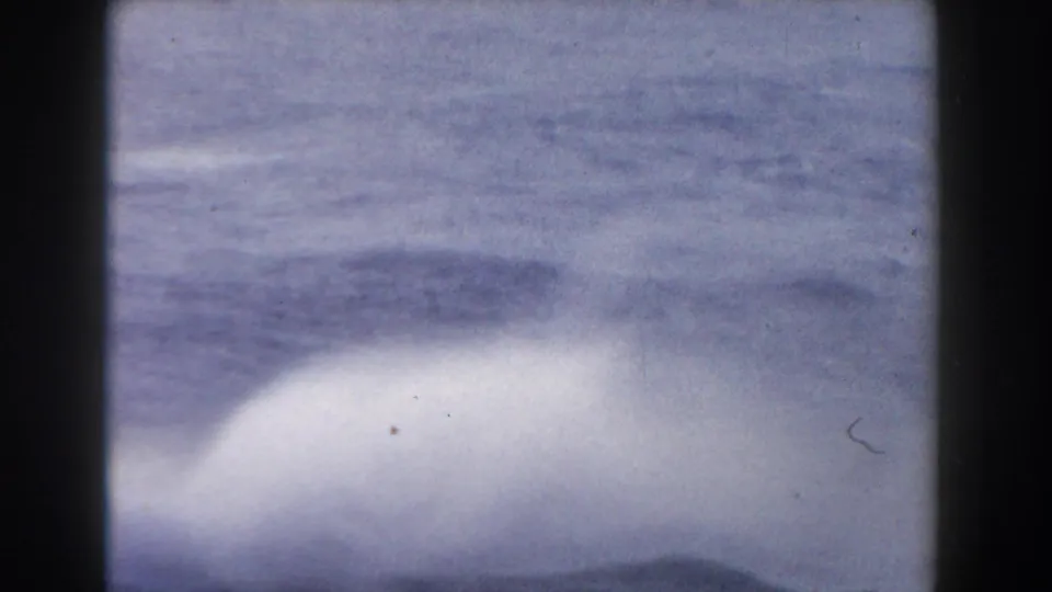Soft, close shipboard view of pale spray and dark blue-gray ocean surface captured on grainy 8mm film.
