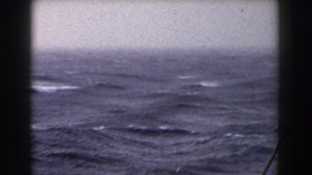 A hazy horizon and rolling blue-gray sea fill the view, partially blocked by dark boat window framing and a thin diagonal line near the right edge.