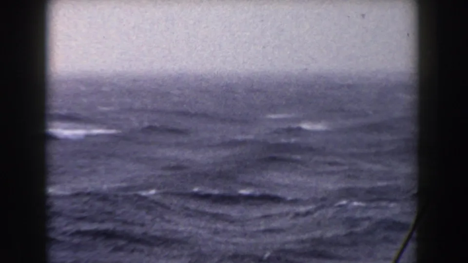 A hazy horizon and rolling blue-gray sea fill the view, partially blocked by dark boat window framing and a thin diagonal line near the right edge.