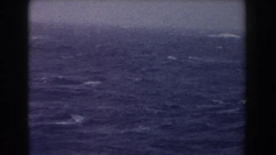 Open Atlantic water appears under a hazy sky, framed by dark shipboard verticals in a faded 8mm home-movie view.