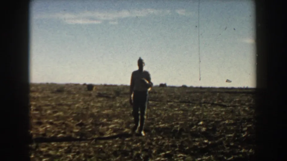A silhouetted man stands in an open sunlit field in 1950s 8mm home-movie footage with a low horizon and pale sky.