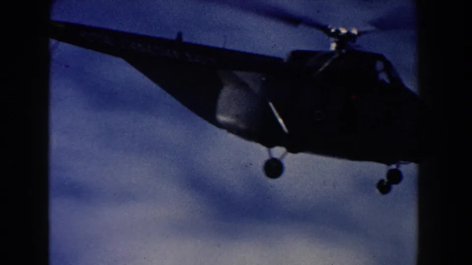 1960 8mm home-movie frame showing a dark helicopter silhouette with blurred rotor blades against a cloudy sky.