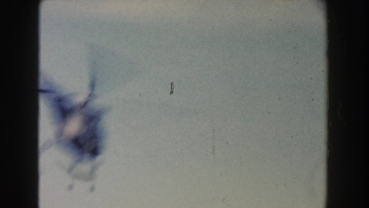 1960 Dearborn home-movie frame showing a pale bluish field, a smeared dark shape at left, and small dust or scratch marks.