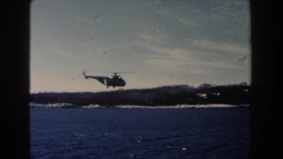 A helicopter approaches a shoreline over dark water in the 1960 Dearborn, Michigan 8mm home-movie footage.