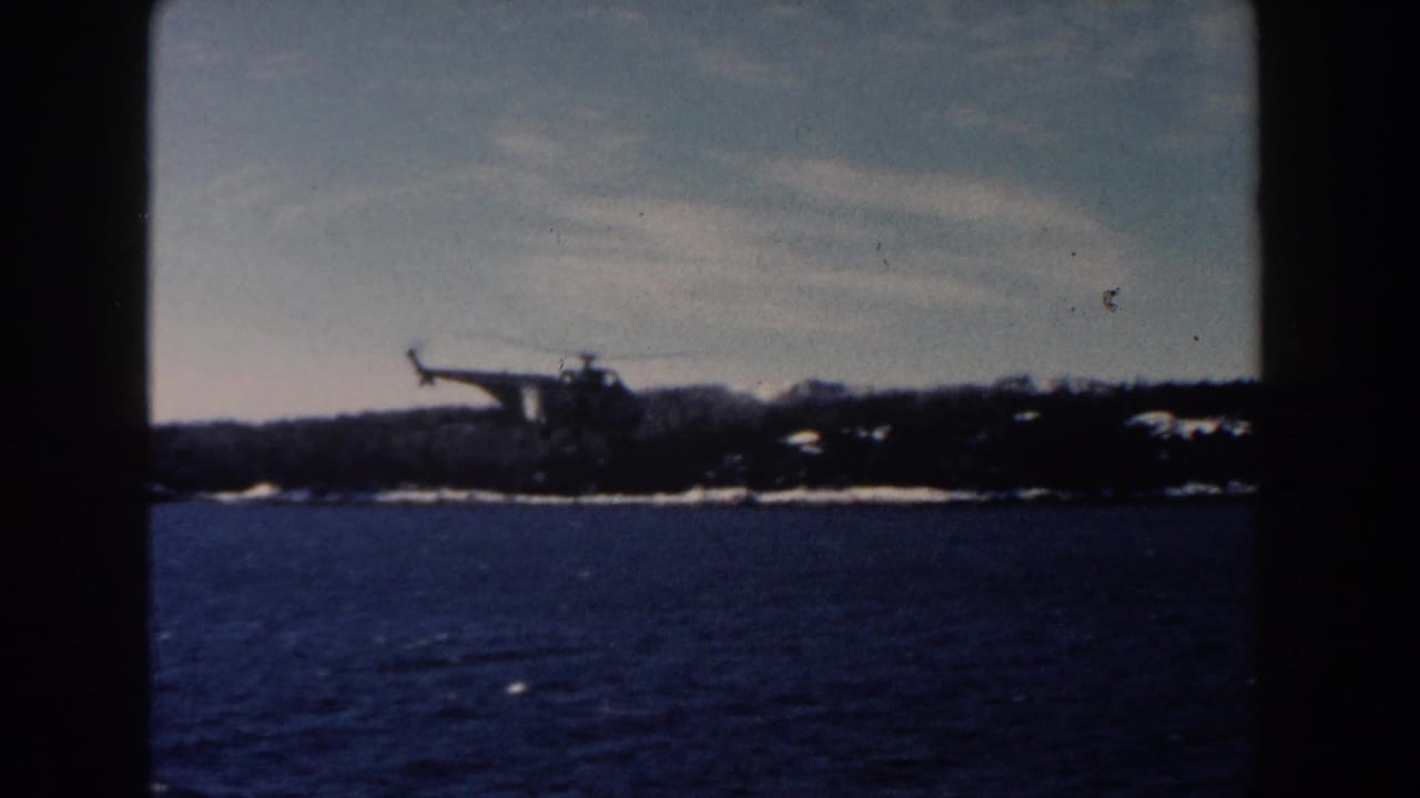 Helicopter flying low over dark water with snowy shoreline in the distance.
