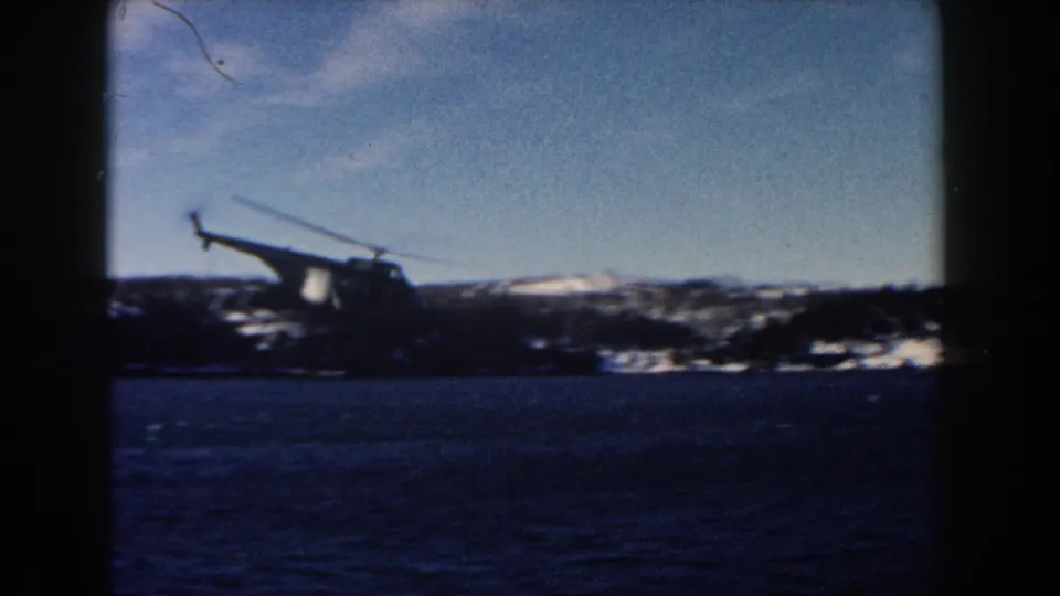 Soft 8mm frame of a helicopter moving over blue water, backed by low snow-covered land and a distant dark horizon.