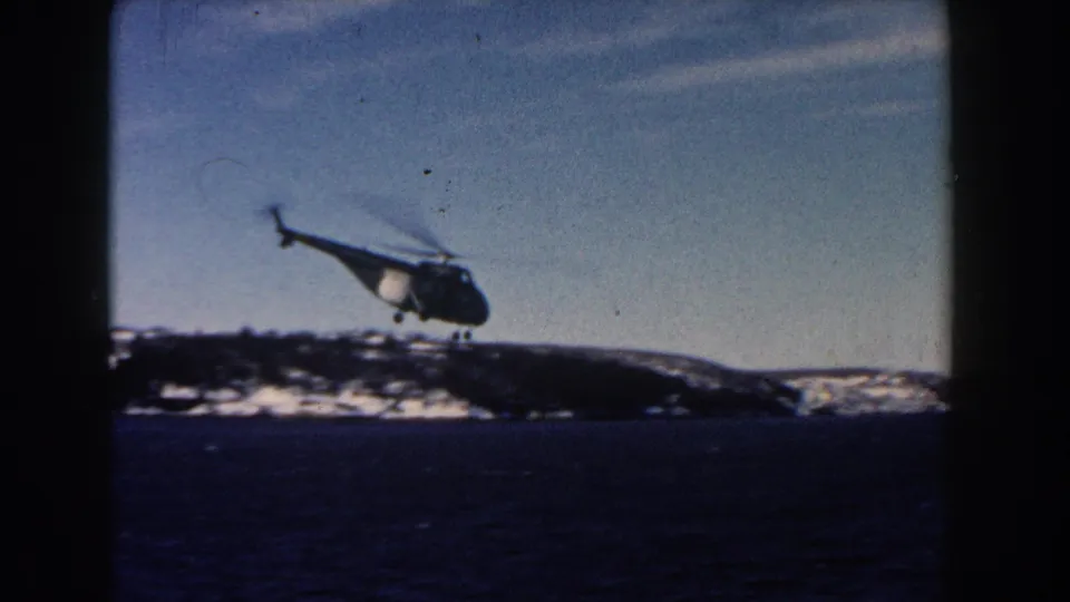 1960 8mm frame showing the helicopter low over a winter waterfront, with dark water in front and a pale sky behind.