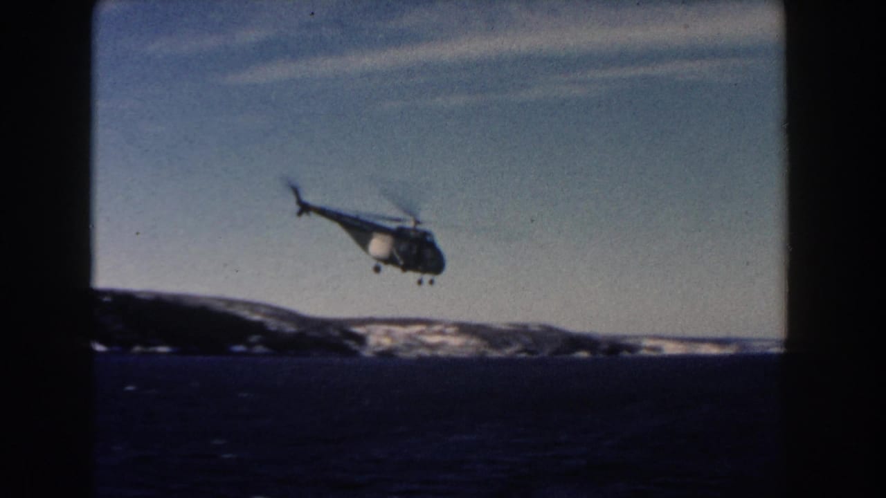 1960 8mm frame showing a small helicopter over open water with snow-dusted land on the horizon.
