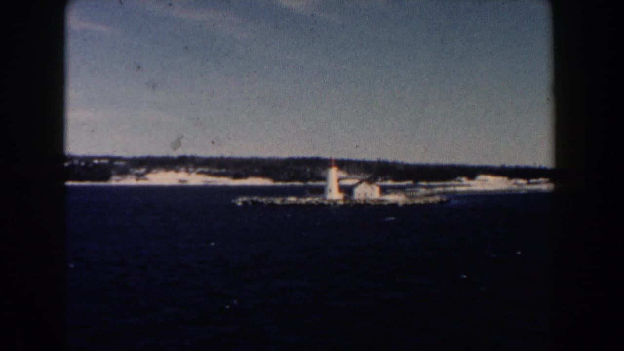 Distant lighthouse and low shoreline seen across dark water in faded 1960 home-movie color.