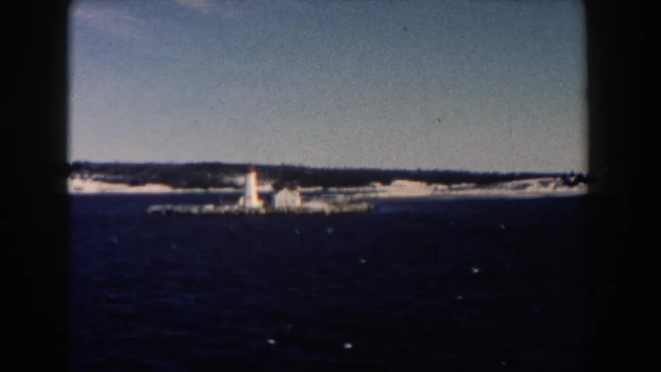 1960 8mm view of a lighthouse, low structures and winter shoreline across open water.
