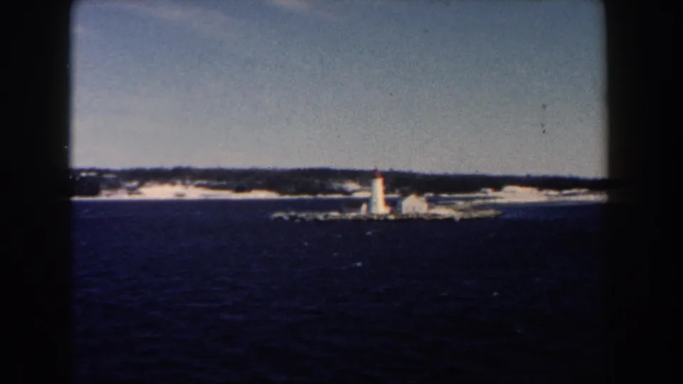 Faded 8mm frame of a white lighthouse and low buildings along a snow-dusted shoreline.