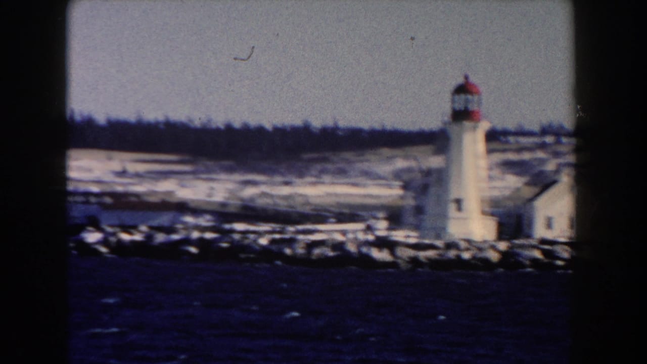 White lighthouse beside snowy shoreline and dark water, seen through 8mm film frame edges.