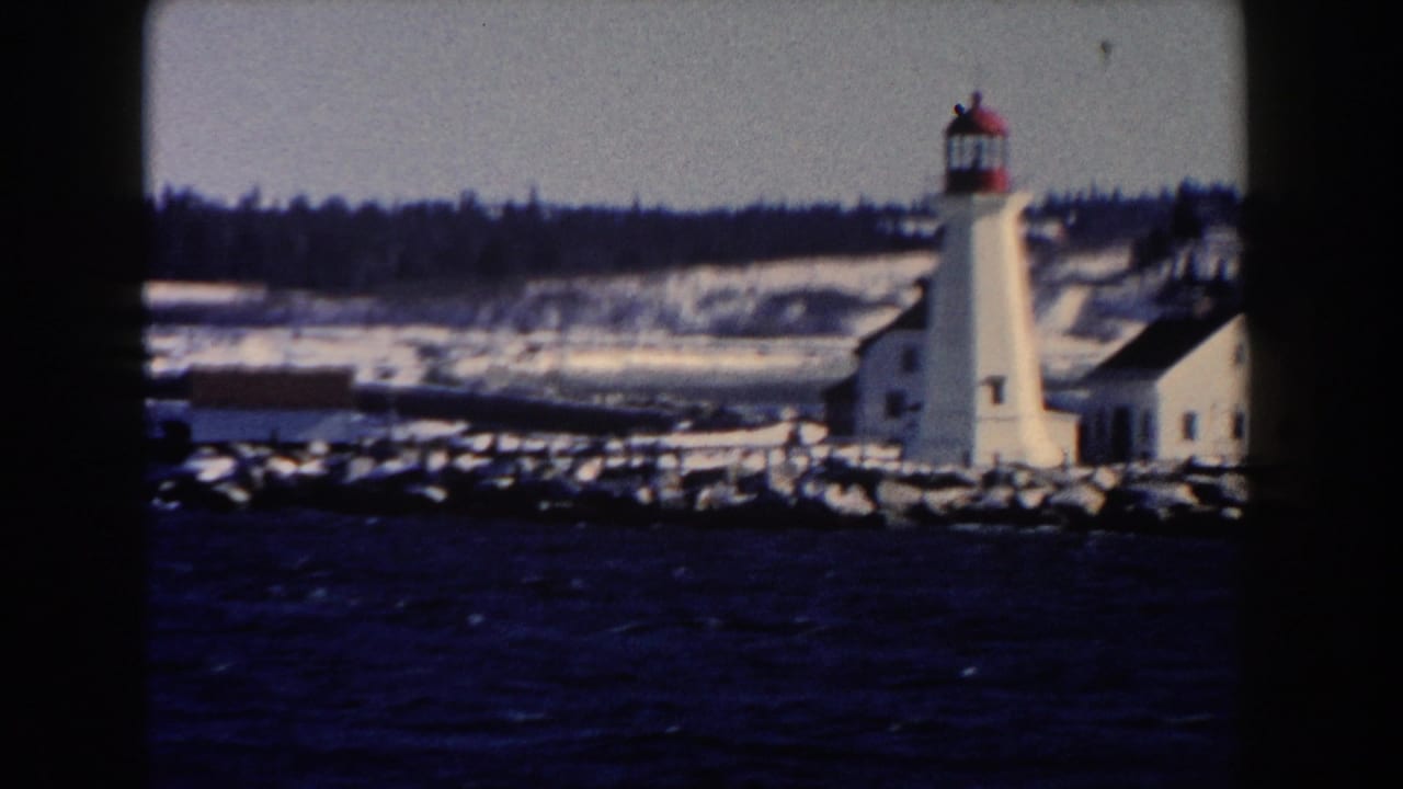 Snowy lighthouse and adjacent building across choppy water with a dark treeline behind.