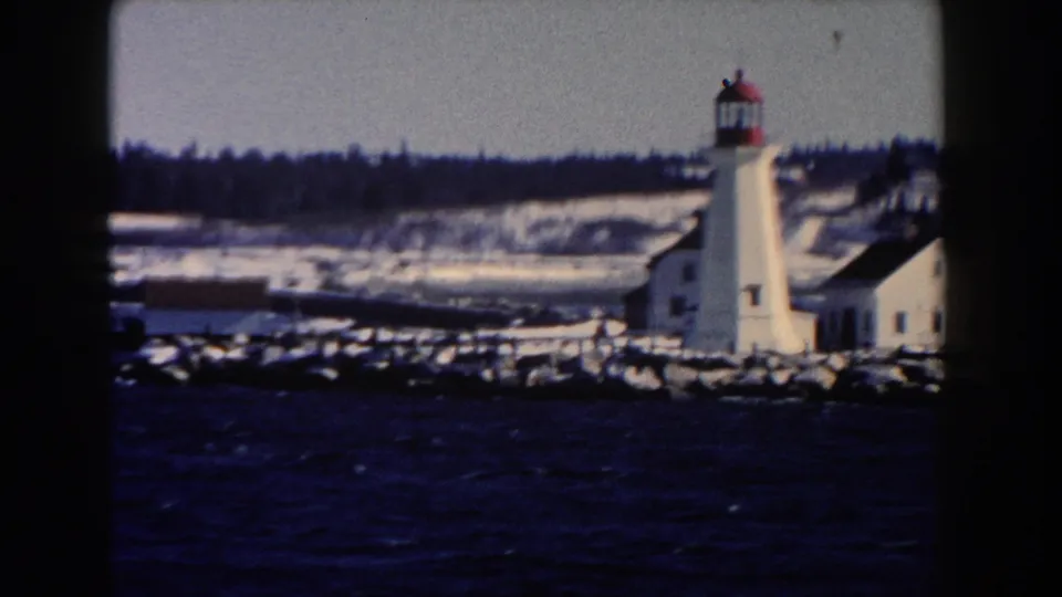 A white lighthouse and nearby low building appear across dark harbor water in the supplied 1960 Michigan home movie.