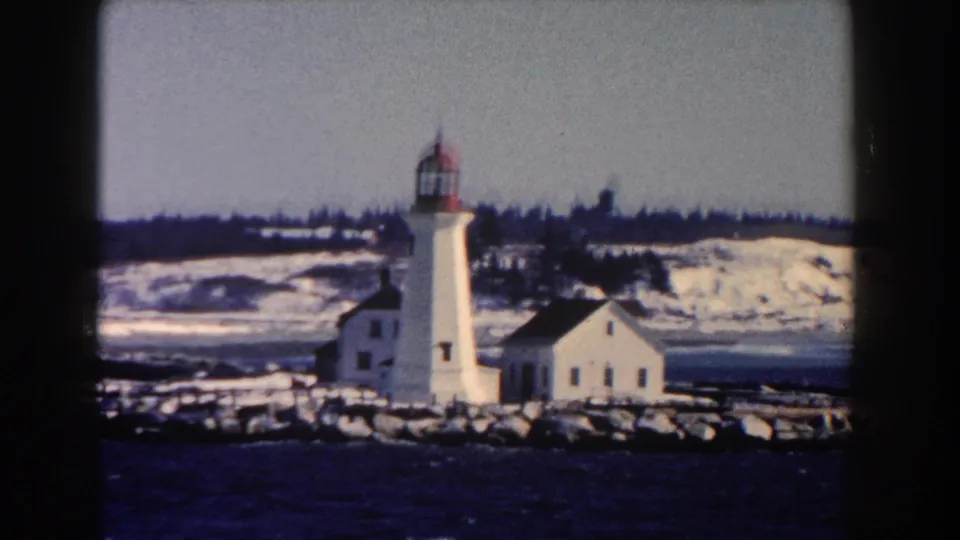 The 1960 8mm footage frames the lighthouse more centrally, with a low building, snow, rocks, and harbor water visible.