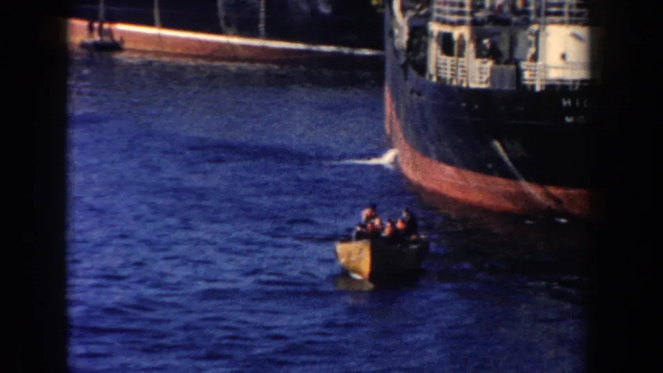 A small boat carrying several people approaches or passes near a large vessel in the 1960 Michigan harbor footage.