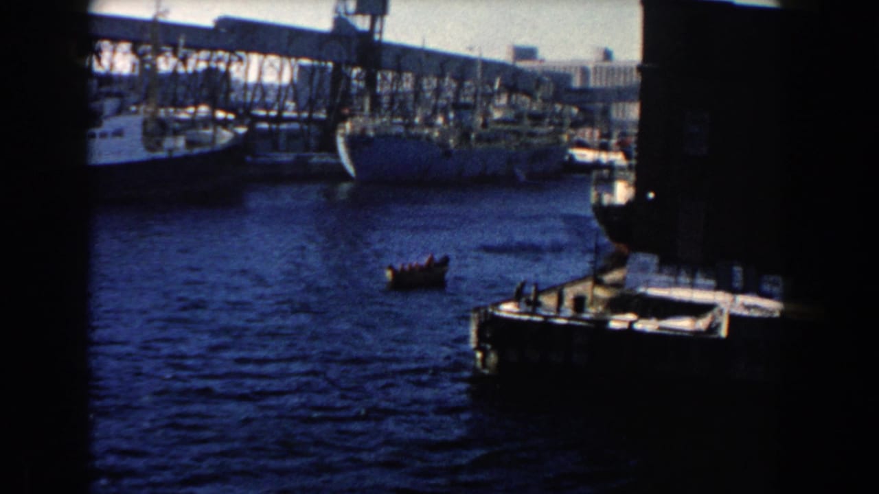 1960 Dearborn harbor view with a small rowboat moving across water near a large cargo ship and industrial docks.