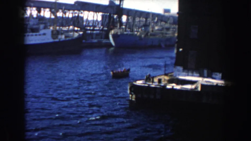 Handheld 8mm frame showing a rowboat near a dock while larger ships line the industrial harbor beyond.