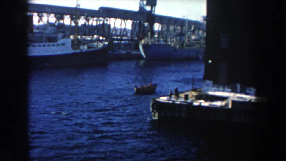 1960 home-movie view of a small rowboat beside dock structures, with large cargo vessels across the harbor.