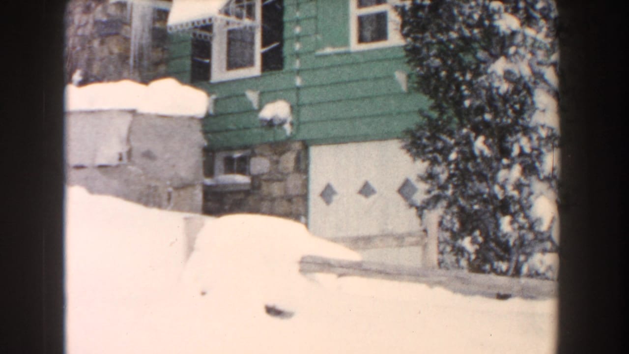 Snow piled against a green-sided house with stone foundation and garage door.