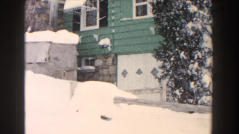 1960 Dearborn, Michigan home-movie frame showing deep snow around a green-sided house with a stone foundation and garage door.