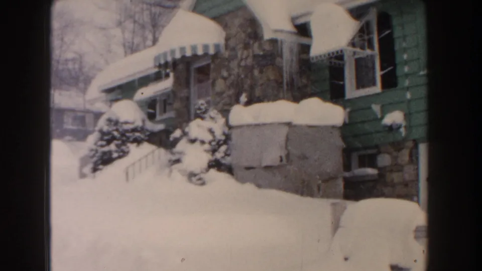 1960 home-movie view of a snowbound Dearborn residence with green siding, stone foundation, evergreen shrubs, and neighboring house visible.