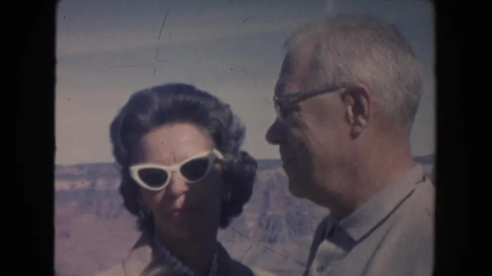 At the canyon overlook, the woman faces the camera in white sunglasses while the man turns toward her in profile.