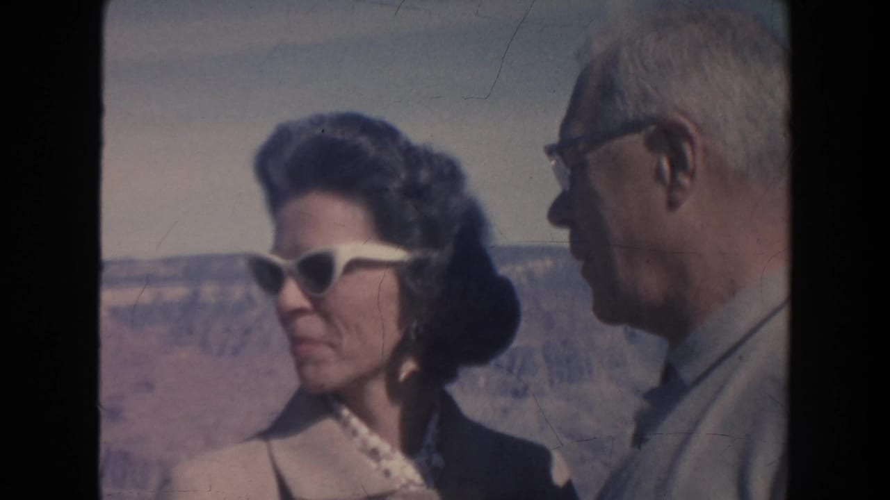1962 Arizona home-movie frame showing a woman in white cat-eye sunglasses speaking with an older man near the Grand Canyon.