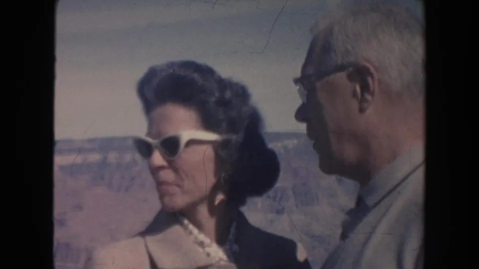 1962 Arizona home-movie frame showing a woman in white cat-eye sunglasses speaking with an older man near the Grand Canyon.
