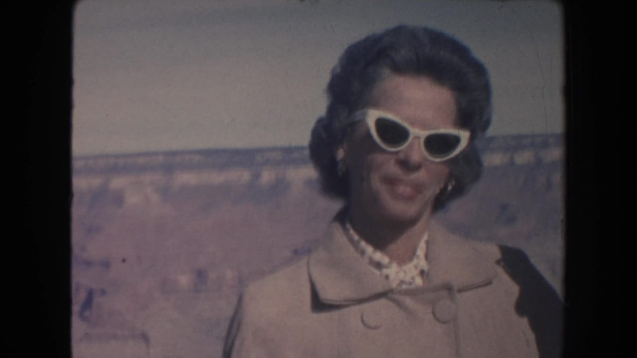 A woman in a light coat and white cat-eye sunglasses smiles at the camera at the Grand Canyon in 1962 home-movie footage.