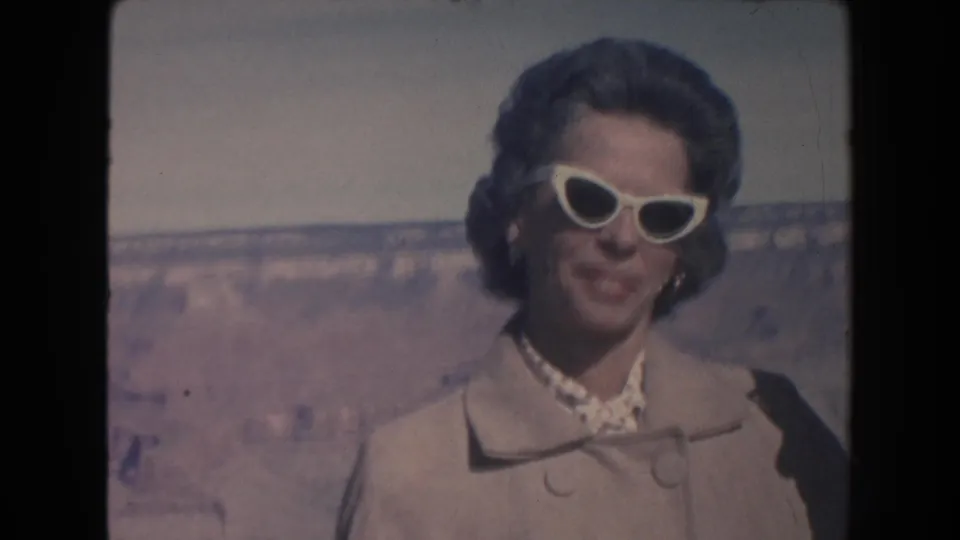 A woman in a light coat and white cat-eye sunglasses smiles at the camera at the Grand Canyon in 1962 home-movie footage.