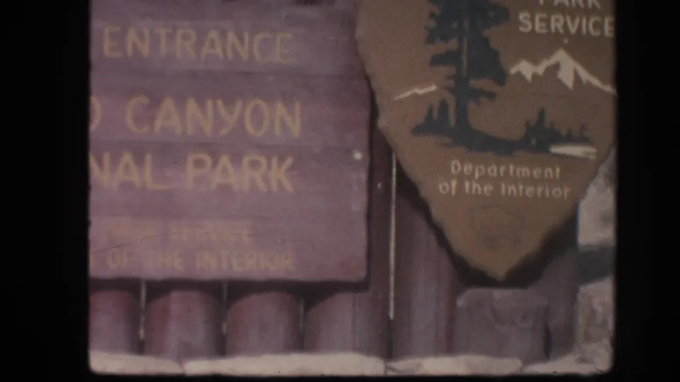 Home-movie close-up of partial Grand Canyon entrance lettering beside the NPS arrowhead in Arizona.