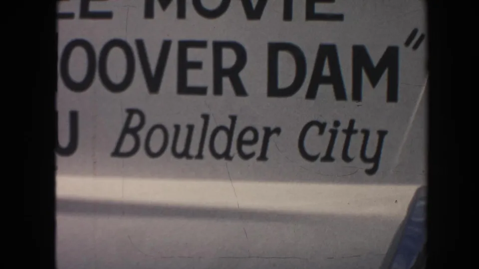 Cropped 8mm frame of the Boulder City Hoover Dam movie-ad sign, with large black lettering and visible film wear.