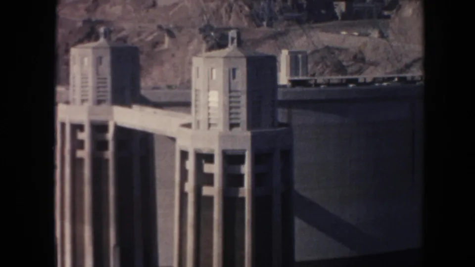 1962 8mm view of Hoover Dam intake towers, concrete dam structure, and desert rock slope in strong sunlight.