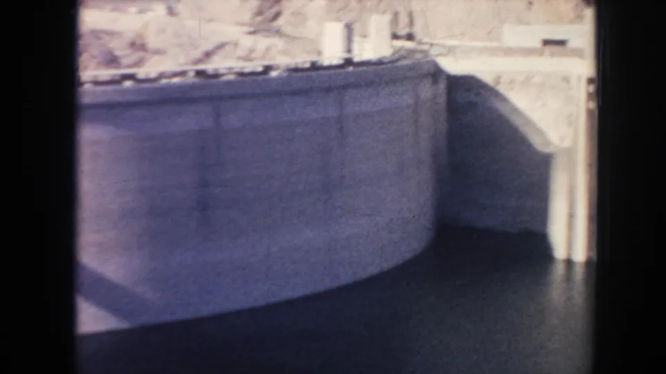 1962 Hoover Dam view of a curved concrete wall, dark reservoir water, rocky terrain, and a bright vertical tower edge.