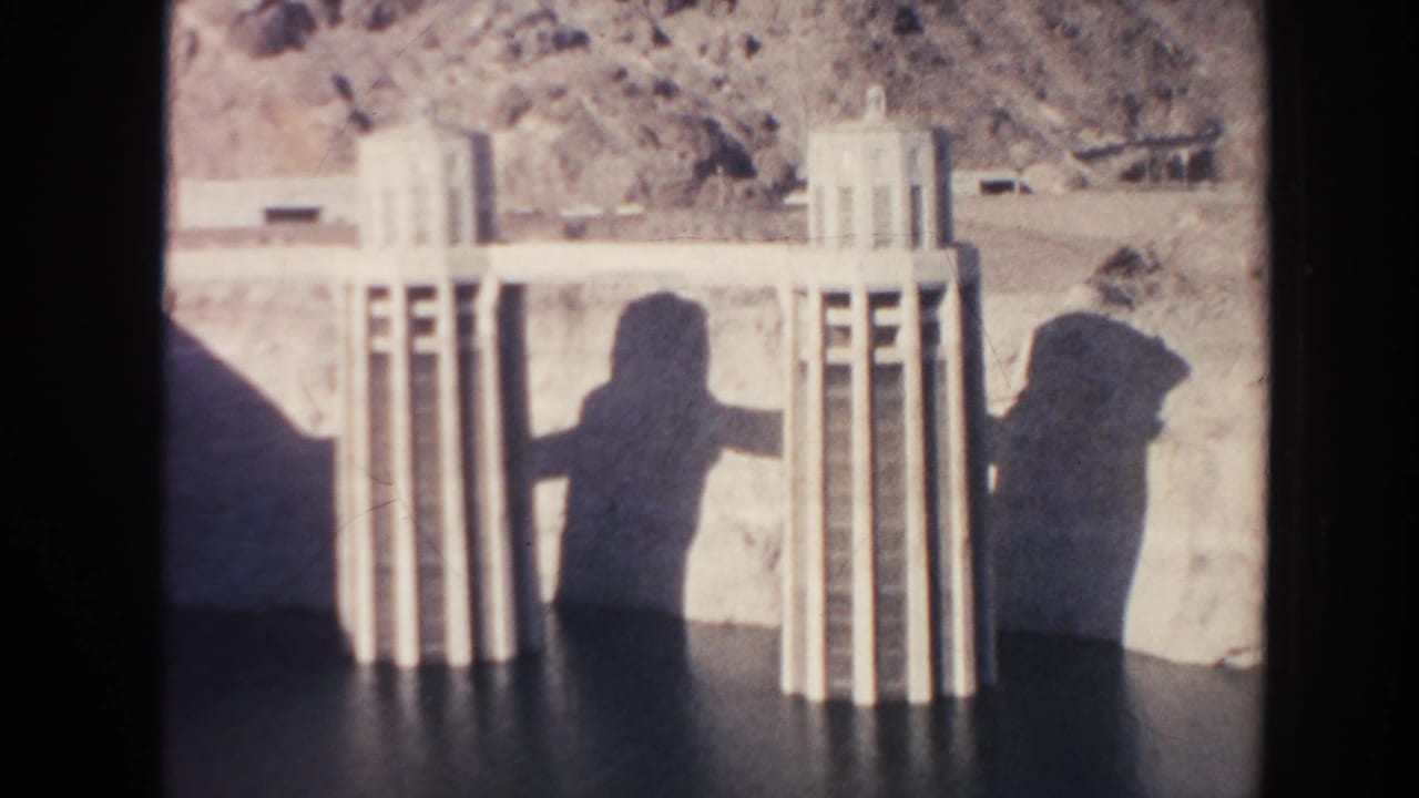 Concrete dam towers beside dark water with two long human shadows.