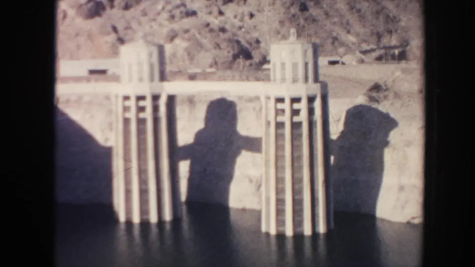1962 8mm frame of concrete dam structures in an Arizona desert setting, with two visitor shadows cast across the water or spillway surface.