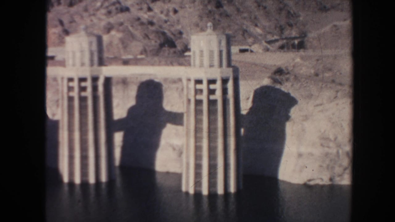 Concrete dam towers and shadowed figures near a rocky desert backdrop.