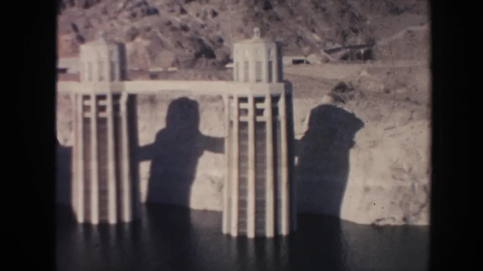 Concrete intake or spillway towers appear against arid hills, with visitor shadows visible on the dam surface in this 1962 Arizona home-movie frame.