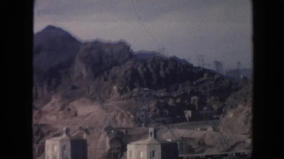 A wide 8mm view shows rugged Arizona desert mountains rising above concrete dam buildings and a dark water or spillway area.