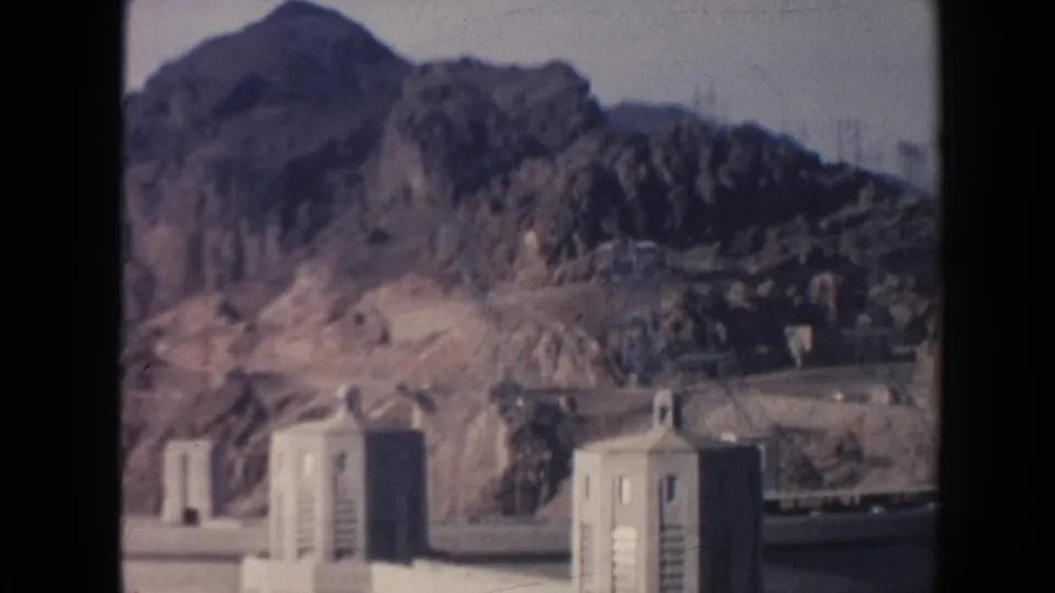 Concrete dam buildings sit below steep arid mountains, with faint utility structures visible in the distance in this 1962 Arizona 8mm frame.