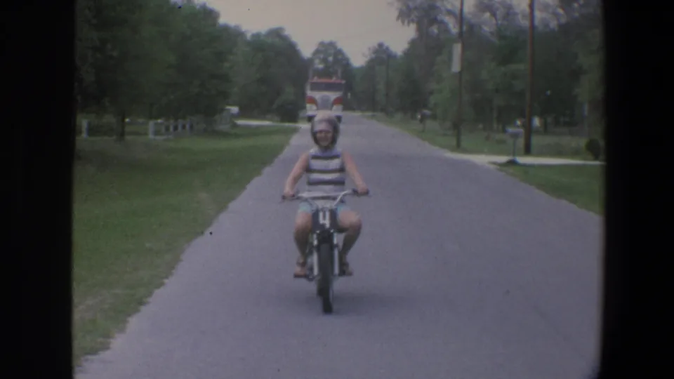 The rider comes nearer on the small motorbike, with grass, trees, and a distant red-and-white truck behind her in Minnesota footage.