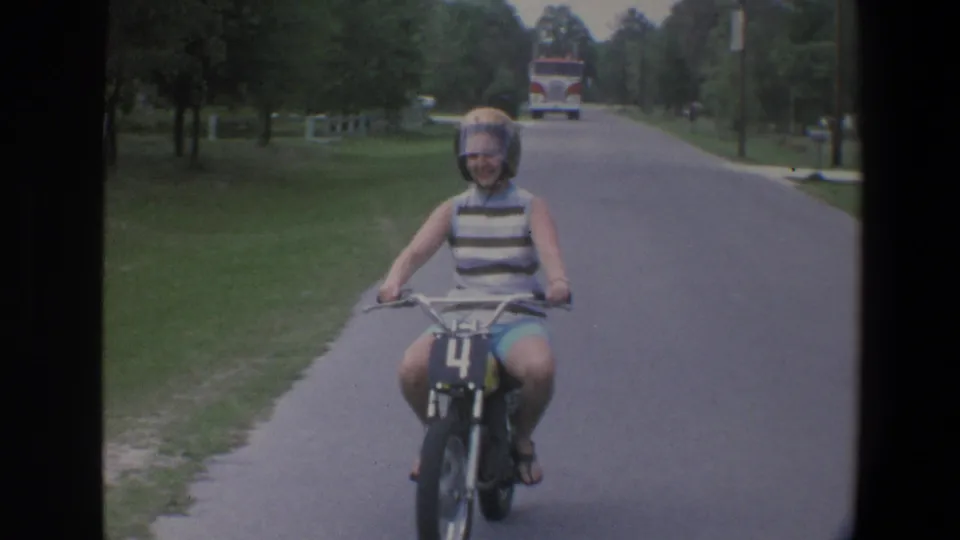 The young woman smiles while riding the small motorbike toward the camera on a green, tree-lined Minnesota road in 1973 footage.