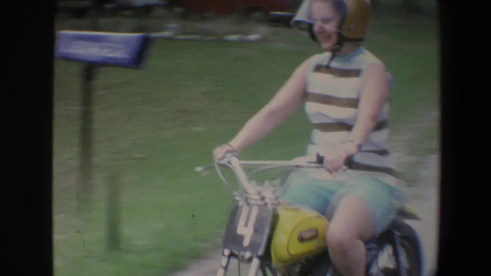A close view shows the smiling rider passing the camera on a small yellow motorbike, with motion blur and green roadside behind her.