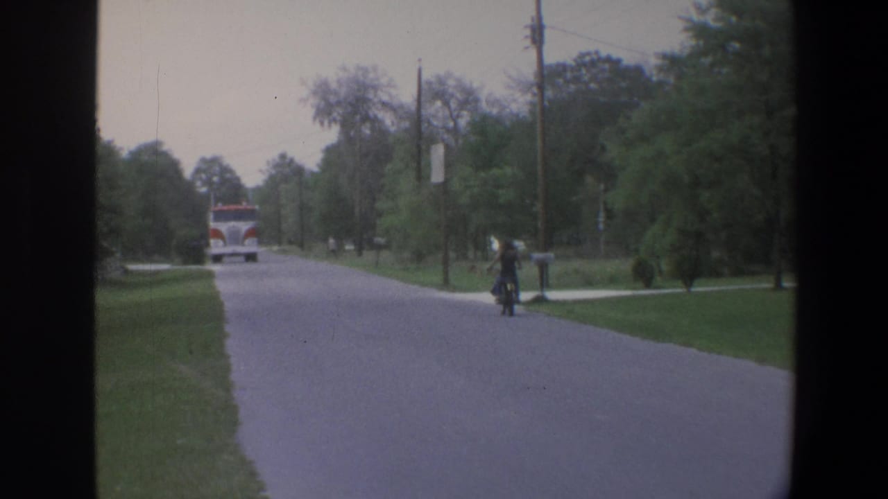 1973 Minnesota 8mm frame of a quiet street lined with lawns, trees, and utility poles, with a red-and-white fire truck in the distance.